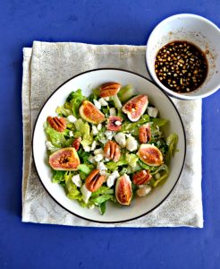 A large bowl filled with mixed greens and topped with ripe pink figs, crumbled white cheese, and pecan halves with a small bowl of balsamic vinaigrette in the upper right hand corner on a sparkly white background.