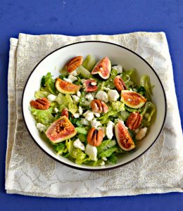 A large bowl filled with mixed greens and topped with ripe pink figs, crumbled white cheese, and pecan halves on a sparkly white background.