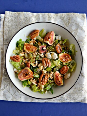 A large bowl filled with mixed greens and topped with ripe pink figs, crumbled white cheese, and pecan halves drizzled with balsamic vinaigrette on a sparkly white background.