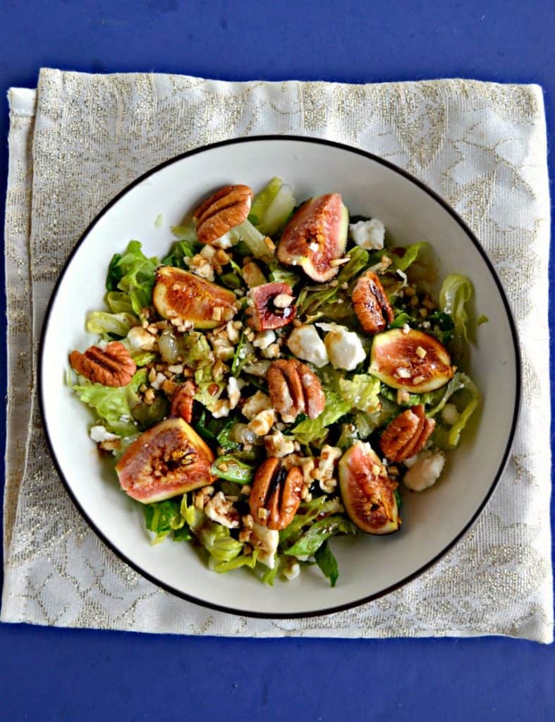 A large bowl filled with mixed greens and topped with ripe pink figs, crumbled white cheese, and pecan halves drizzled with balsamic vinaigrette on a sparkly white background.
