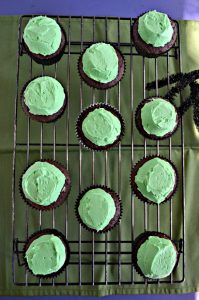 A green background with a wire cooling rack on it and 12 chocolate cupcakes topped with green frostin gand a sider peeking in from the right.