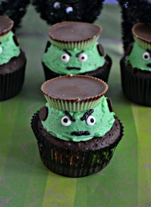 Two Frankenstein Cupcake sitting one behind the other with green frosting, angry eyebrows, edible google eyes, and a peanut butter hat sitting on a green striped background.