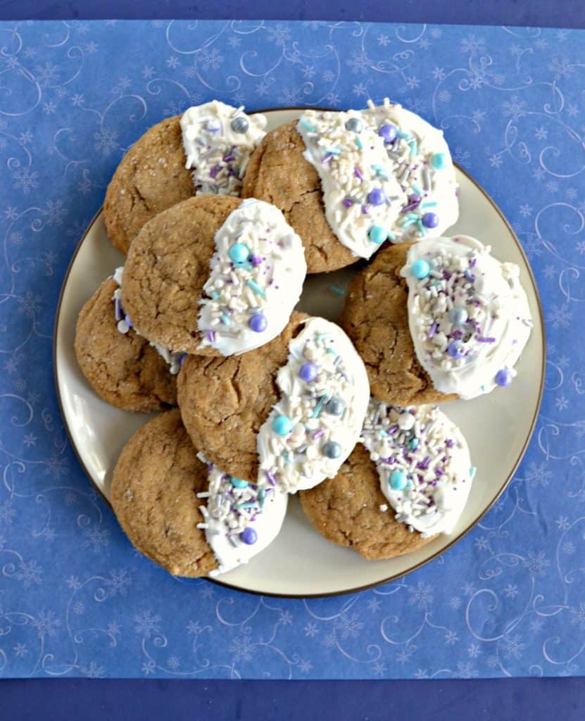 A white plate piled high with brown ginger cookies that are all dipped in white chocolate on the right half of the cookies and sprinkled with white, silver, and blue sprinkles on a blue background.