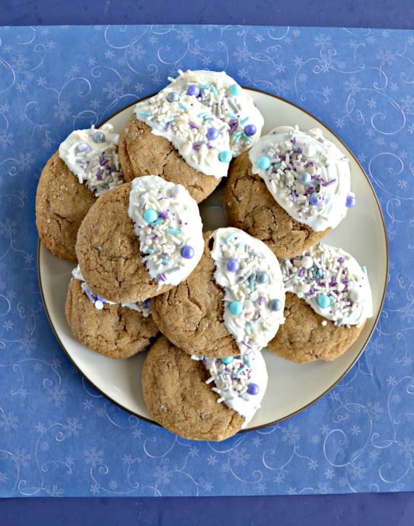 A white plate piled high with brown ginger cookies that are all dipped in white chocolate on the right half of the cookies and sprinkled with white, silver, and blue sprinkles on a blue background.