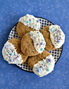 A blue and white checked plate topped with 5 brown ginger cookies arranged on the plate, each one half dipped in white chocolate with white, purple, and blue sprinkles on a blue background.