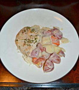 A white plate with a pile in the middle that is onions, red skin potatoes, and carrots with a pork chops on top all in a white gravy.