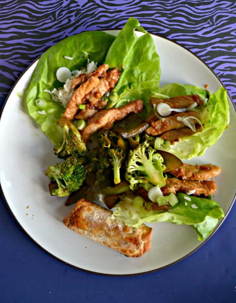 A close up view of a white place on a blue background. There are three lettuce leaves on the plate, each filled with rice and strips of pork with a mound of broccoli on top and an egg roll on the bottom portion of the plate.
