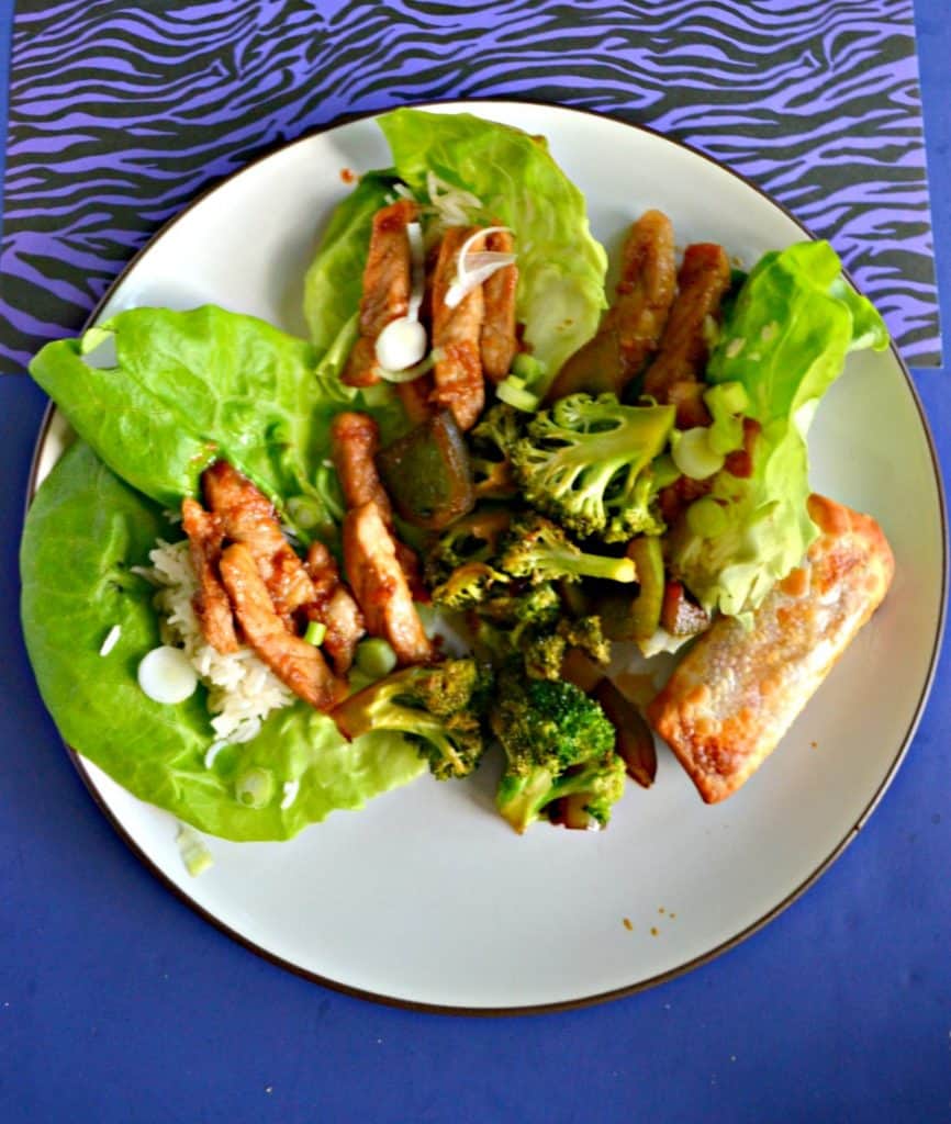 A white plate sitting on a blue backgound. The plate has three leaves of lettuce each topped with serveral strips of pork with rice peaking out from underneath. There's a mound of broccoli in the center and an eggroll on the bottom right portion of the plate.