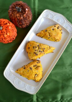A green background with a white platter laying diagonal and 3 orange colored scones on the platter studded with brown chocolate chips and in the upper left hand corner is a glittery orange pumpkin and a glittery brown pumpkin.