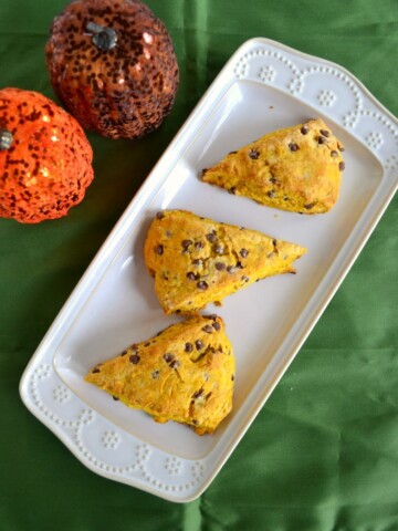 A green background with a white platter laying diagonal and 3 orange colored scones on the platter studded with brown chocolate chips and in the upper left hand corner is a glittery orange pumpkin and a glittery brown pumpkin.