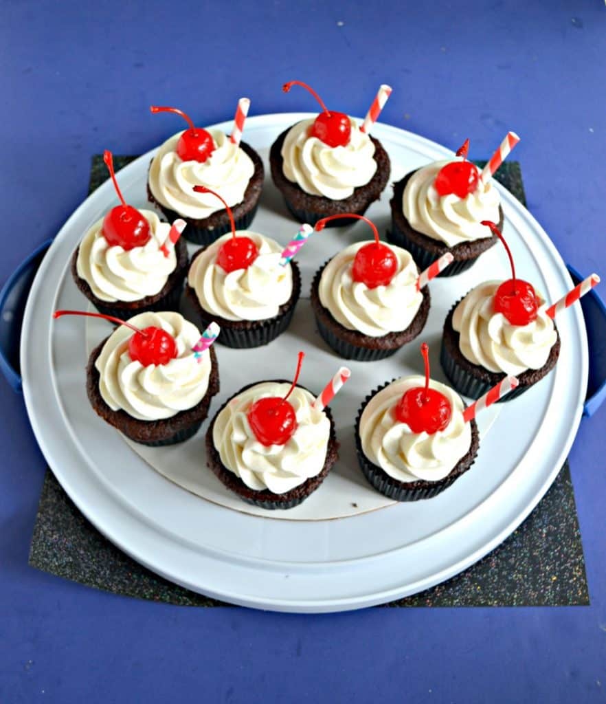 A white, round platter topped with a dozen chocolate cupcakes topped with white frosting, a red cherry, and a straw sticking out of it on a blue background.