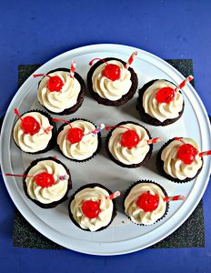 A top view of a white, round platter topped with a dozen chocolate cupcakes topped with white frosting, a red cherry, and a straw sticking out of it on a blue background.