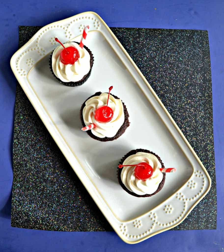 A top view of a white platter with three chocolate cupcakes on it with white frosting and a cherry on top on a sparkly black background.