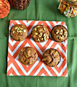 Two rows of muffins, the bottom row has two muffins, the top row has three muffins topped with pepitass and they are on an orange and white place mat on a green background. There are two glitter pumpkin on the top left and a squat scarecrow on the top right.
