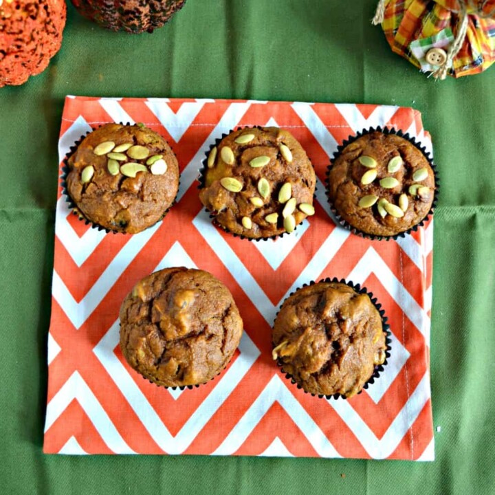 Two rows of muffins, the bottom row has two muffins, the top row has three muffins topped with pepitass and they are on an orange and white place mat on a green background. There are two glitter pumpkin on the top left and a squat scarecrow on the top right.