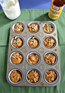 A muffin tin filled with pumpkin batter. The top 6 are topped with pepitas. On the top right is an empty can of pumpkin and on the top left in a jar of sourdough starter.