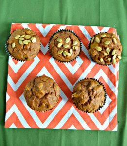 A top view of two rows of muffins, the bottom row has two muffins, the back row has three muffins topped with pepitass and they are on an orange and white place mat on a green background.