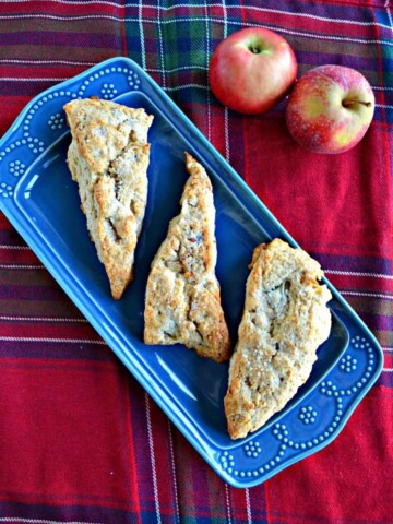 A blue platter with three large, golden brown scones on it on a red plaid background with two apples in the back right corner.