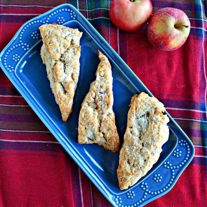 A blue platter with three large, golden brown scones on it on a red plaid background with two apples in the back right corner.