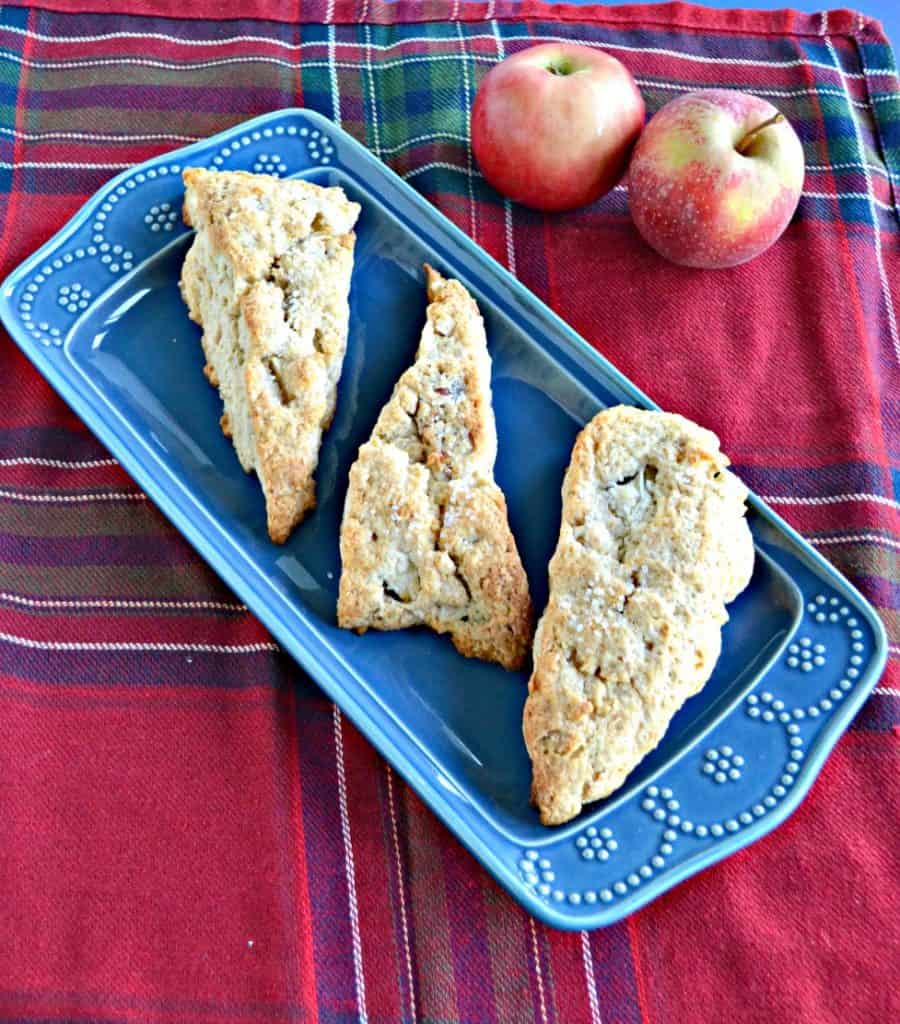 A blue platter with three large, golden brown scones on it on a red plaid background with two apples in the back right corner. 