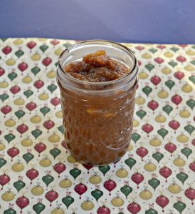 A glass jar filled with brown apple butter on a yellow background with yellow, red, and green ornaments on it.