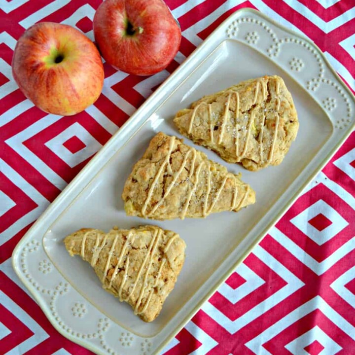 A top view of a white platter with 3 triangular scones on the plate drizzled with caramel glaze on a red and white background and two red apples in the upper left hand corner.