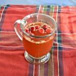 A clear mug filled with red liquid and cranberries floating on top on a red and green plaid tablecloth.