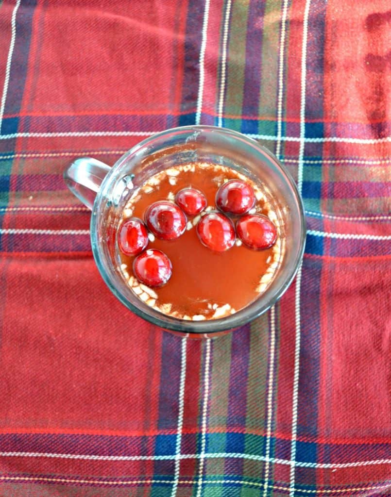 A top few of a clear mug with red liquid in it and cranberries floating on top sitting on a red and green plaid tablecloth.