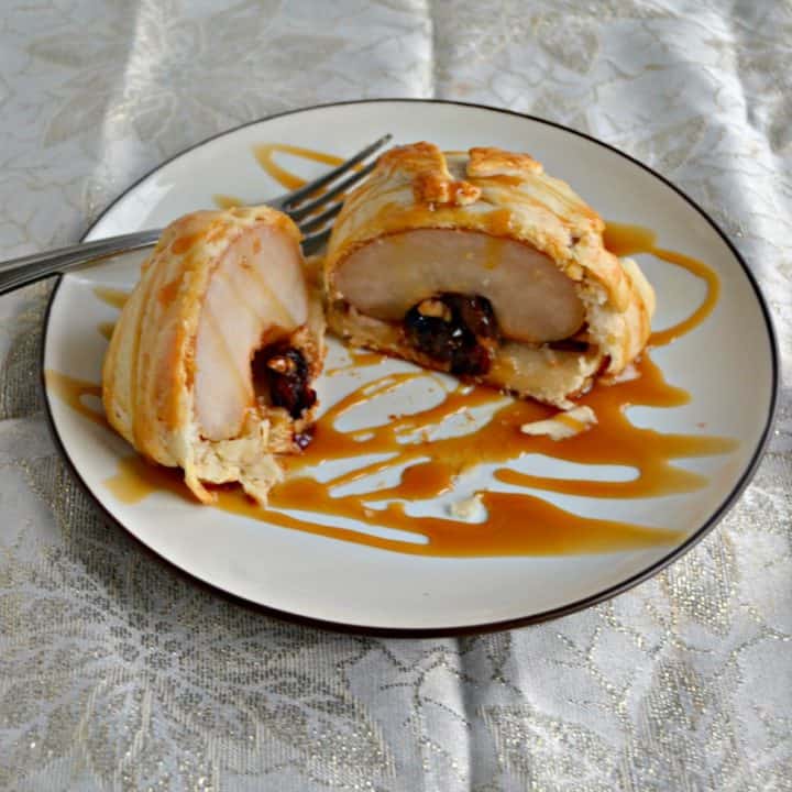 A plate with a pie crust covered pear that is cut in half and at the bottom is stuffed with pecans, chocolate, and cranberries with a fork behind the pear and caramel drizzled over top of it.