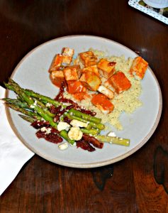 A plate with couscous on the upper right corner and bright orange tofu with asparagus in the lower left corner topped with red tomatoes and white feta cheese,