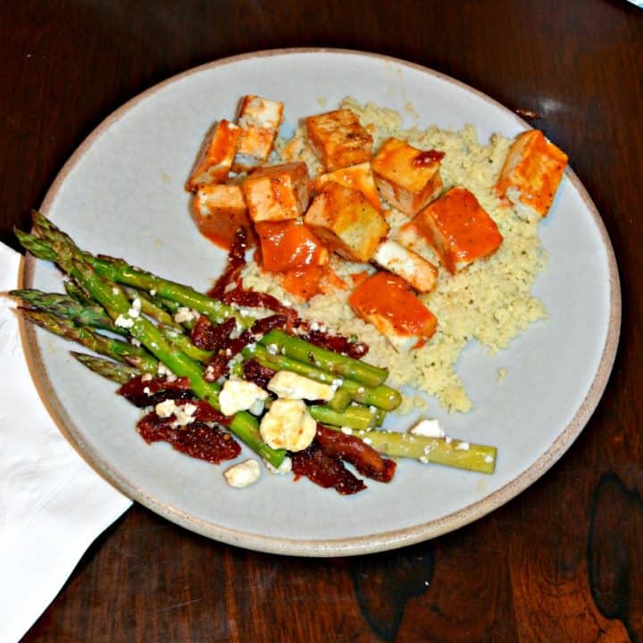 A plate with couscous on the upper right corner and bright orange tofu with asparagus in the lower left corner topped with red tomatoes and white feta cheese,