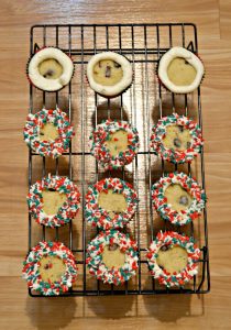 A dozen cupcakes on a wire baking rack. The three cupcakes on the top row have a white frosting ring piped around it, the other nine cupcakes have a ring of sprinkles around them on a wooden background.