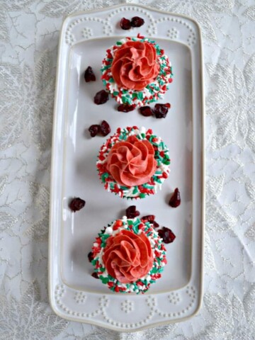 A white platter with three cupcakes on it. Each cupcake has a white ring of frosting with sprinkles on it with red frosting piped in the middle on a white background.