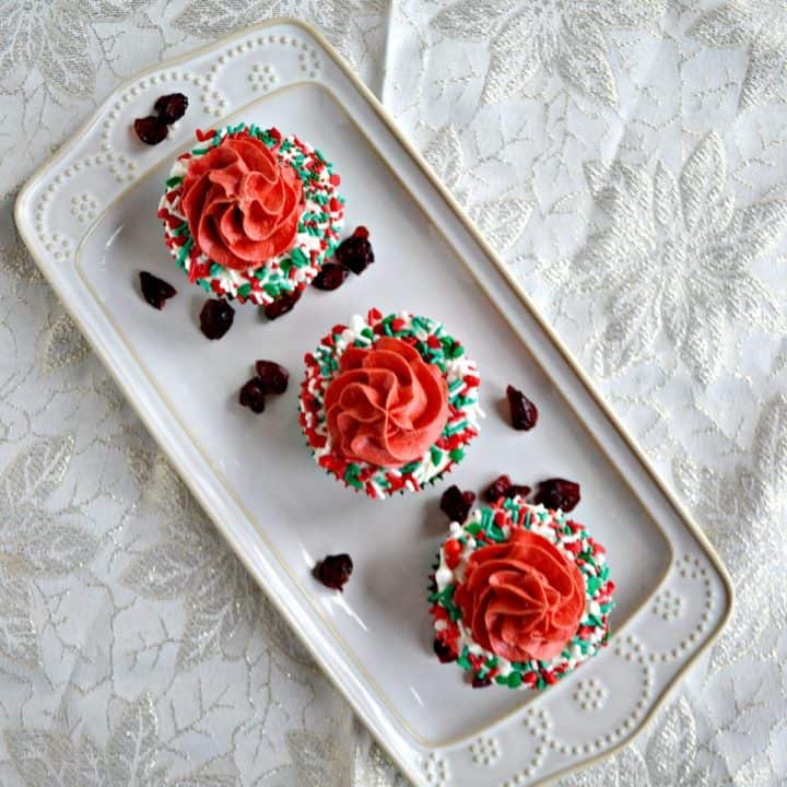 A white platter sitting diagonally with three cupcakes on it. Each cupcake has a white ring of frosting with sprinkles on it with red frosting piped in the middle on a white background.