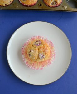 A top view of the golden brown muffin studded with purple grapes and the red and white cupcake liner opened underneath the muffins on a blue background.