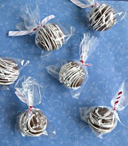 A blue background with six hot chocolate spheres drizzled with white chocolate, wrapped in plastic bags with red and white ribbon tying them, spread out over the blue backdrop.