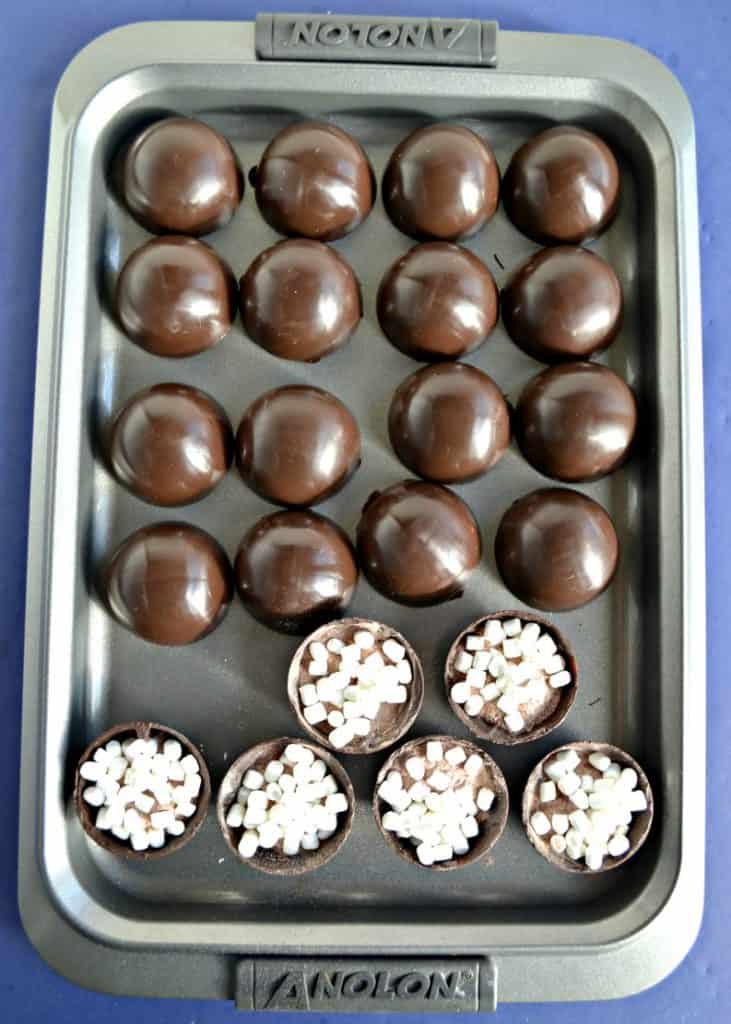 A cookie sheet with rows of chocolate spheres in the back and two rows of half spheres filled with mini marshmallows in the front. 