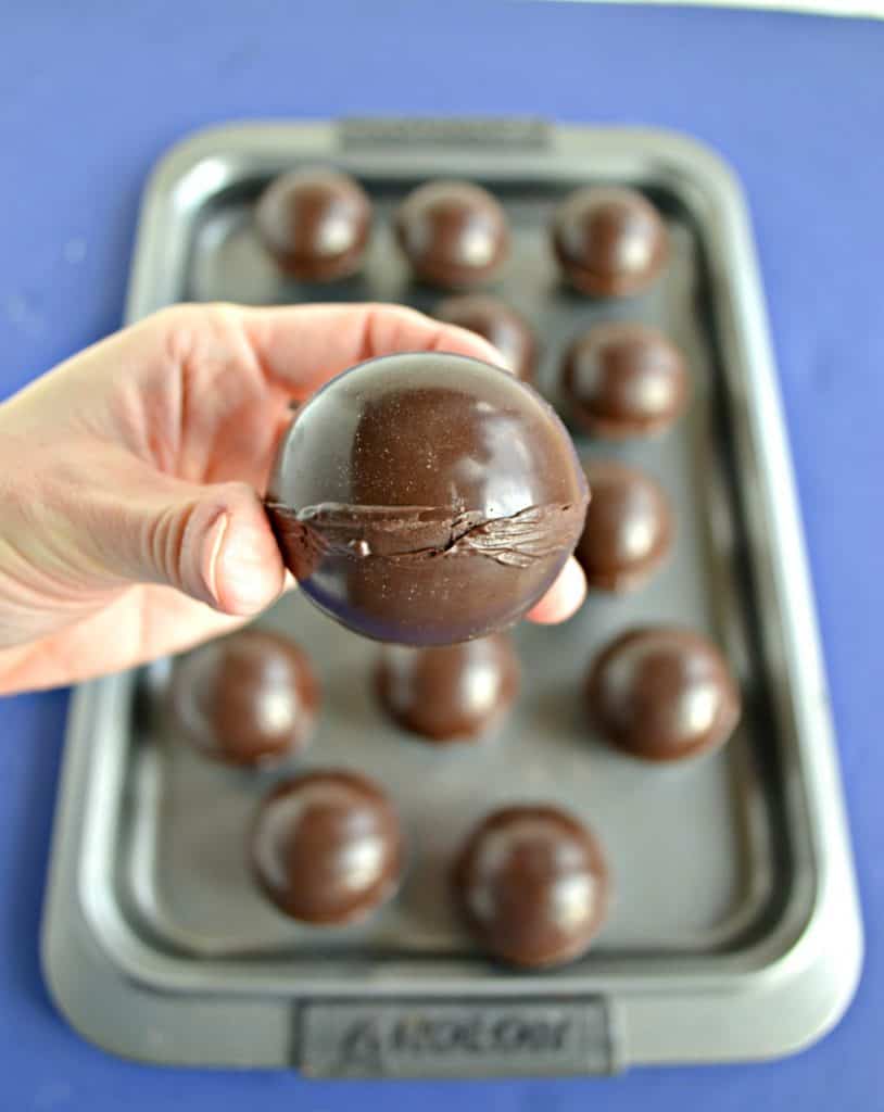 A close up of a hand holding a chocolate sphere with a fuzzy picture of more chocolate spheres on a cookie sheet in the background. 