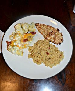 A white dinner plate with a golden brown chicken breast in the upper right hand corner, rice in the lower right hand corner, and a mixture of cauliflower, cheese, a cream sauce, and bacon in the upper left hand corner.