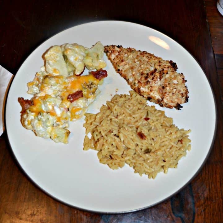 A white dinner plate with a golden brown chicken breast in the upper right hand corner, rice in the lower right hand corner, and a mixture of cauliflower, cheese, a cream sauce, and bacon in the upper left hand corner.
