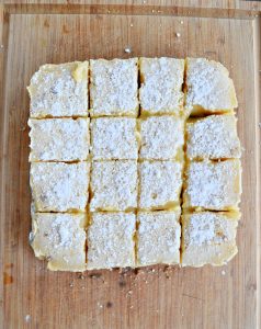 Lemon bars cut into 16 squares topped with powdered sugar and sitting on a brown cutting board.