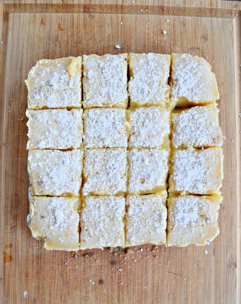 Lemon bars cut into 16 squares topped with powdered sugar and sitting on a brown cutting board. 