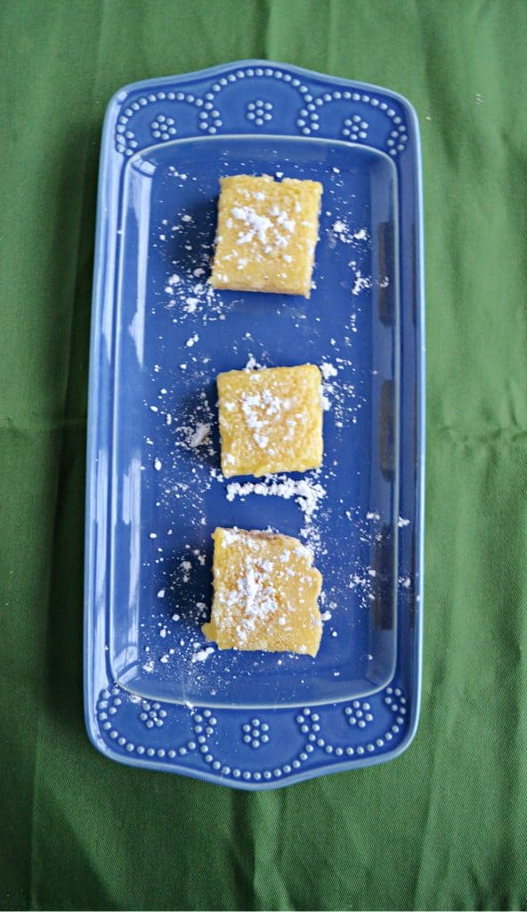 A blue platter sitting vertically with three lemon bars, topped with powdered sugar, sitting on top of it on a green background.