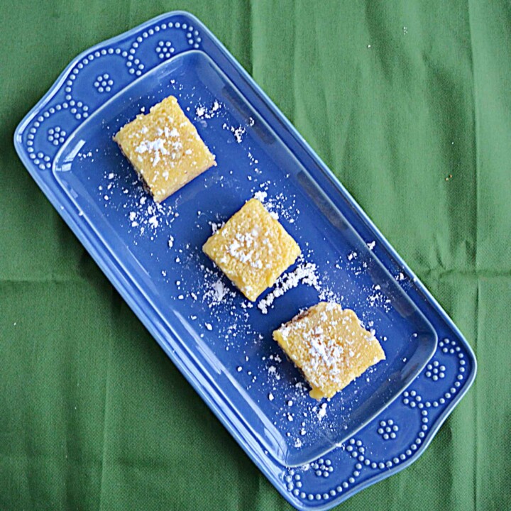 A blue platter sitting diagonally with three lemon bars, topped with powdered sugar, sitting on top of it on a green background.