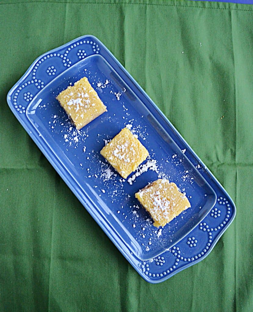 A blue platter sitting diagonally with three lemon bars, topped with powdered sugar, sitting on top of it on a green background.
