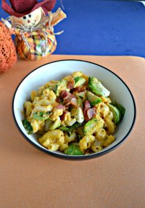 A bowl filled with orange macaroni noodles, green Brussels sprouts, red bacon, all on a sparkling orange background with a stuffed scarecrow in the upper lefthand corner.