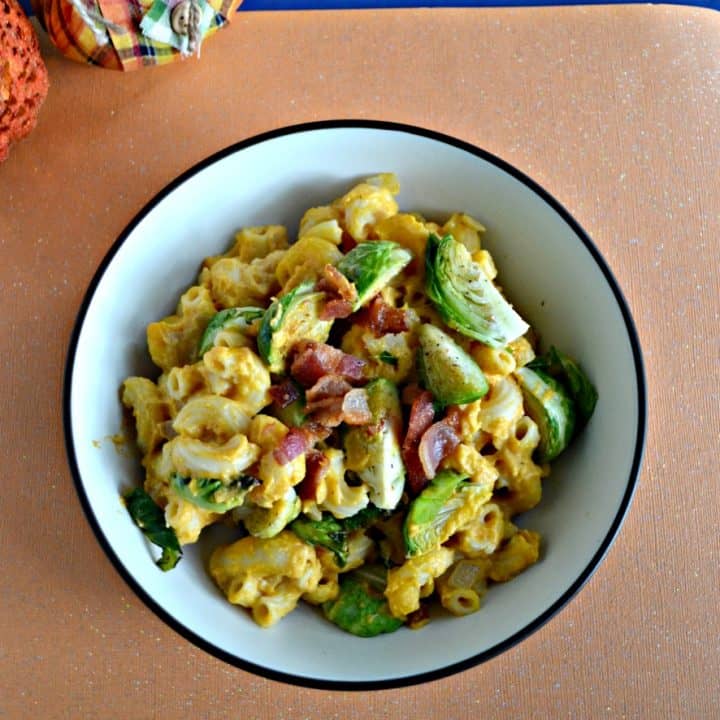 Top view of a bowl filled with orange macaroni noodles, green Brussels sprouts, red bacon, all on a sparkling orange background with a stuffed scarecrow in the upper lefthand corner.