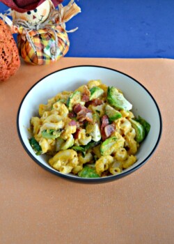 A white bowl sitting on ann orange backgrounf. It is filled with orange macaroni noodles, green Brussels Sprouts, and red bacon with a stuffed pumpkin and stuffed scarecrow in the upper lefthand corner.