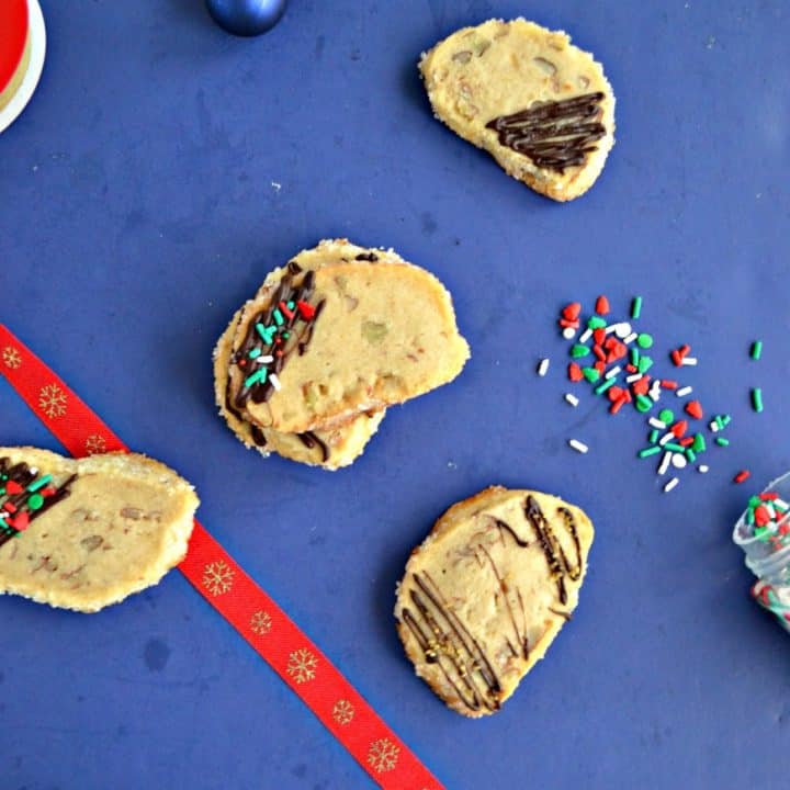 A blue background scattered with shortbread cookies that have been drizzled with chocolate and sprinkled with festive sprinkles. There's a red ribbon down the left side and sprinkles spilled on the right side.