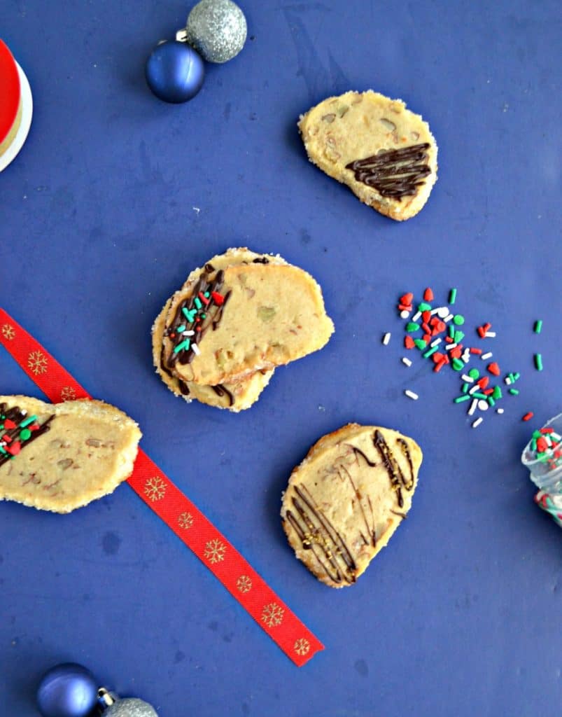 A blue background scattered with shortbread cookies that have been drizzled with chocolate and sprinkled with festive sprinkles. There's a red ribbon down the left side and sprinkles spilled on the right side. 